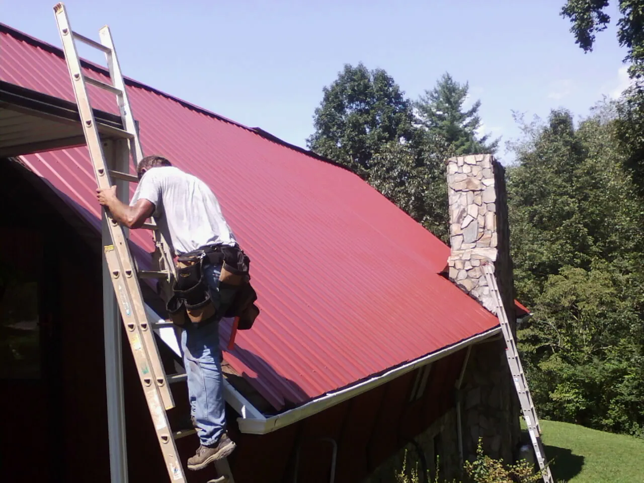 Roofer on a ladder beside a red metal roof and stone chimney — Soffit & Fascia Repair by Roofing Brothers in Greenbank